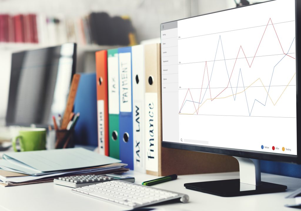 Monitor z wykresem finansowym na biurku z kalkulatorem i kolorowymi segregatorami (TAX LAW, FINANCE).