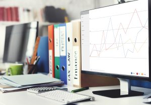 Monitor z wykresem finansowym na biurku z kalkulatorem i kolorowymi segregatorami (TAX LAW, FINANCE).